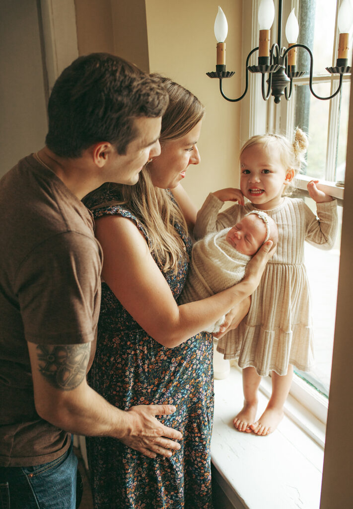 Parents holding their newborn baby girl and a toddler daughter near a small window in Morristown NJ